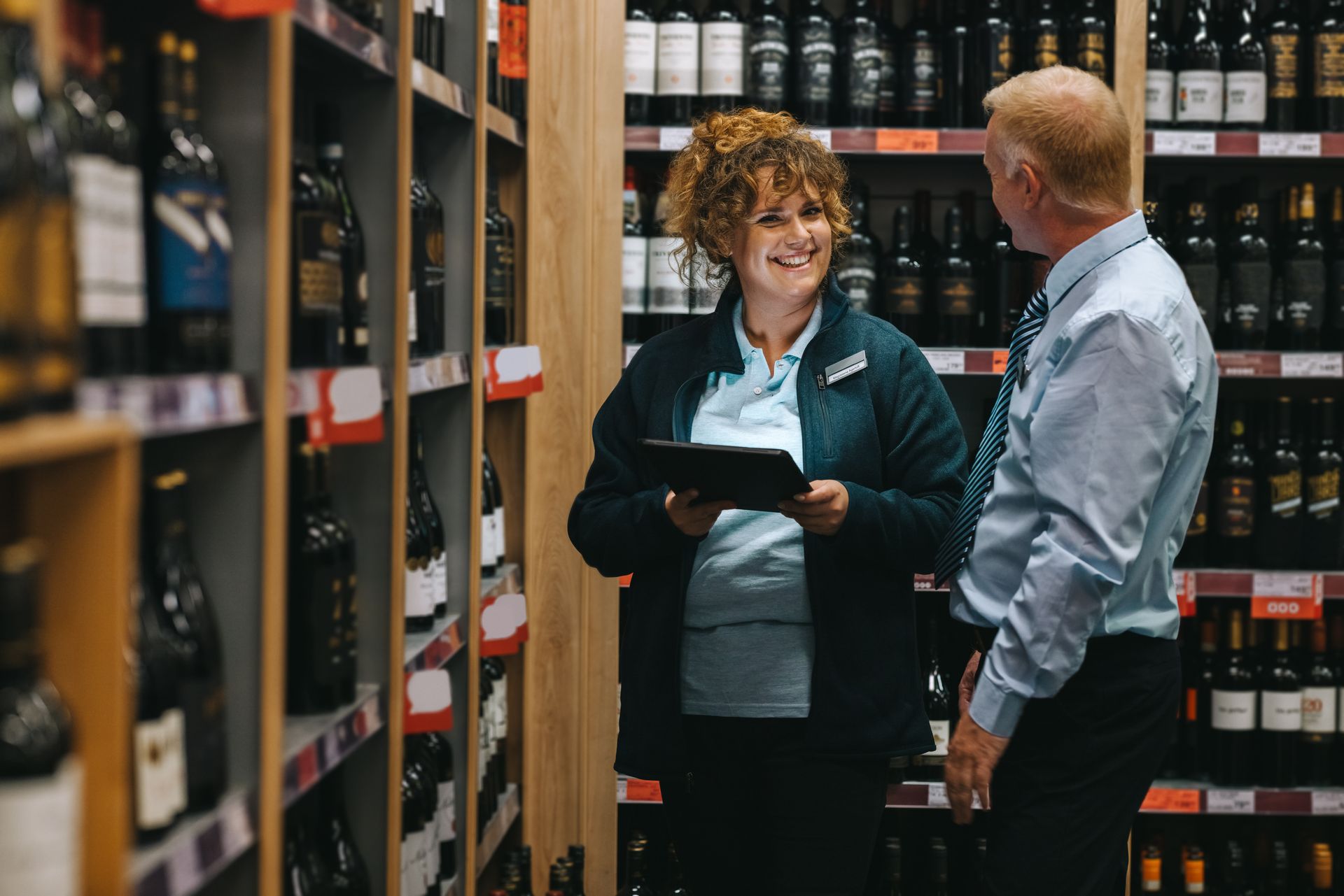 Woman in store uniform smiling, talking to a man near shelves of wine.