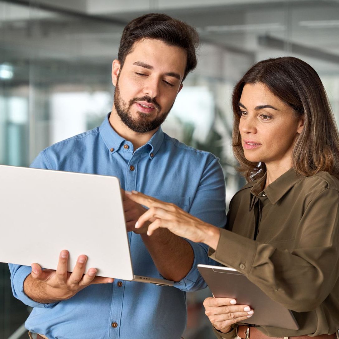 Man and woman collaborating over a laptop and tablet in a modern office setting.
