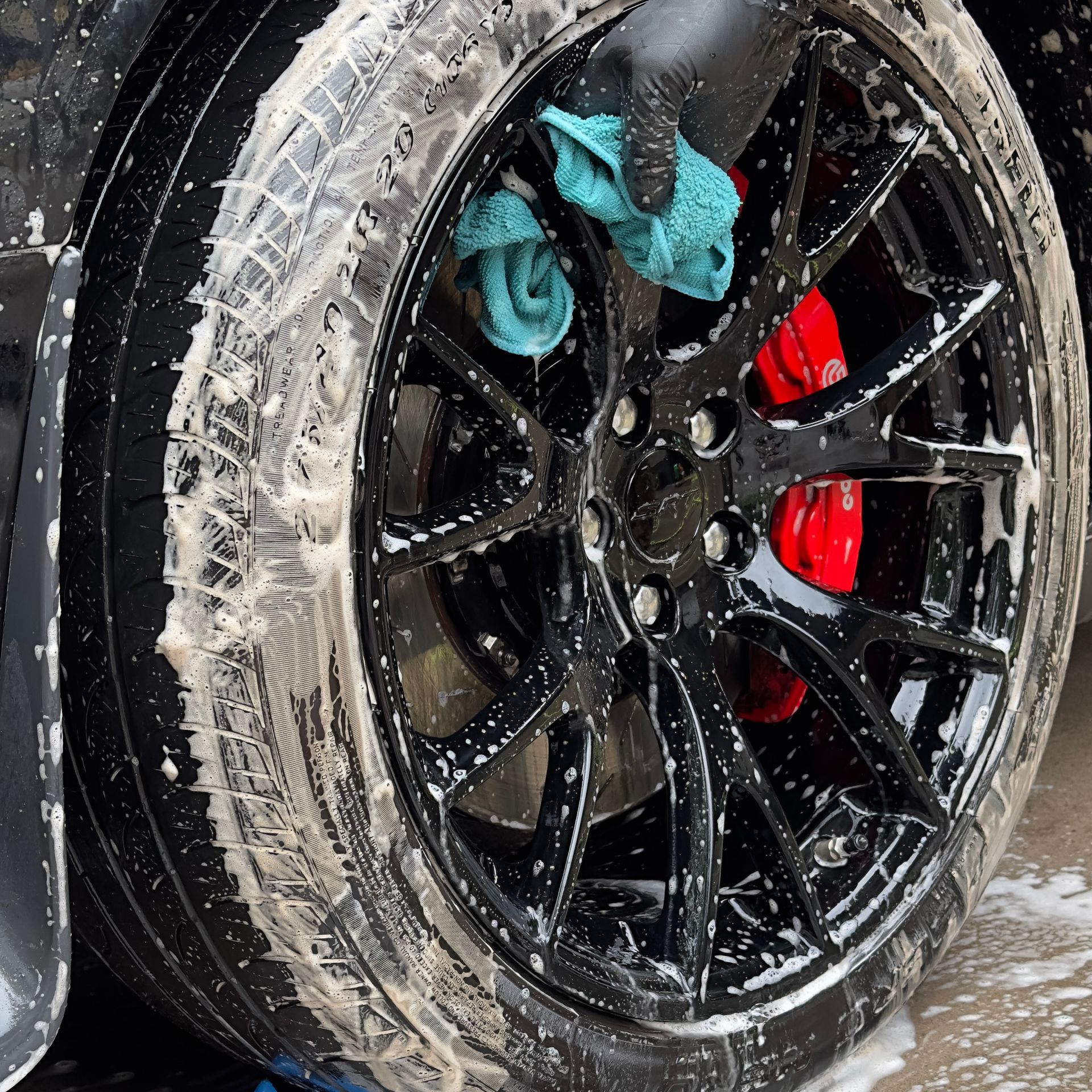 Black car wheel being cleaned with soap and a cloth; red brake calipers visible.