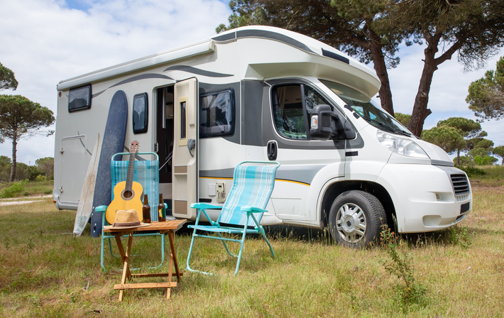A rv is parked in a grassy field with chairs , a guitar , and a surfboard.