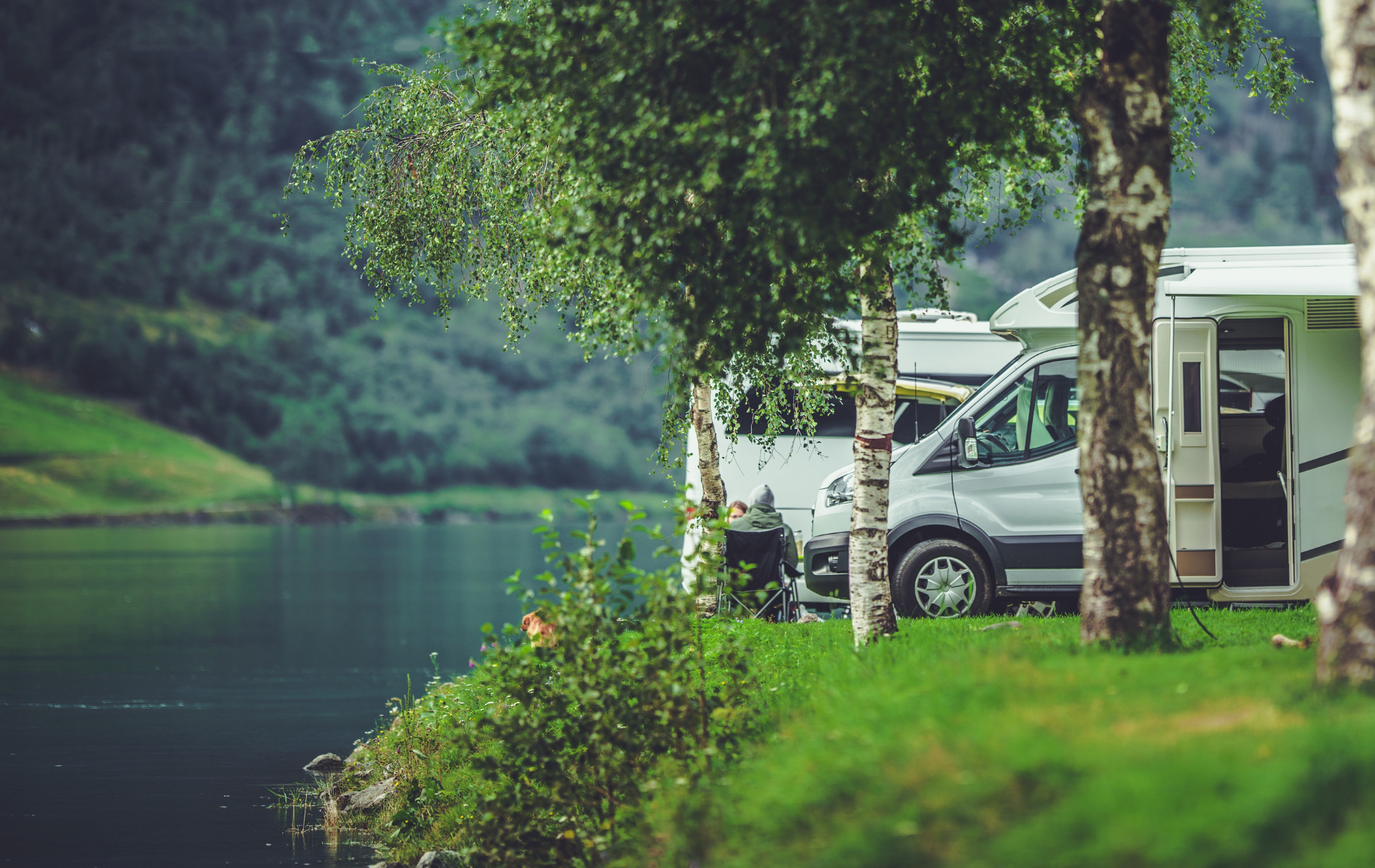 RV camping by a lake, with mountains in the background. Green grass and trees frame the scene.
