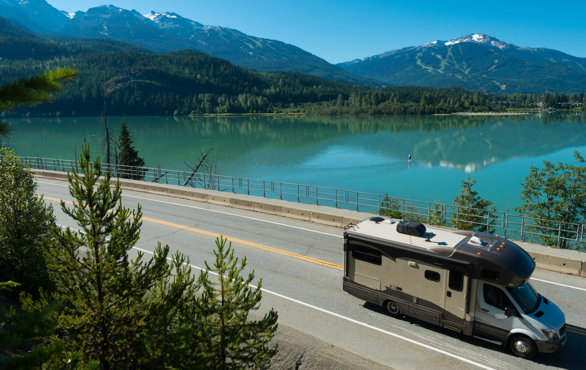 A rv is driving down a road next to a lake with mountains in the background.
