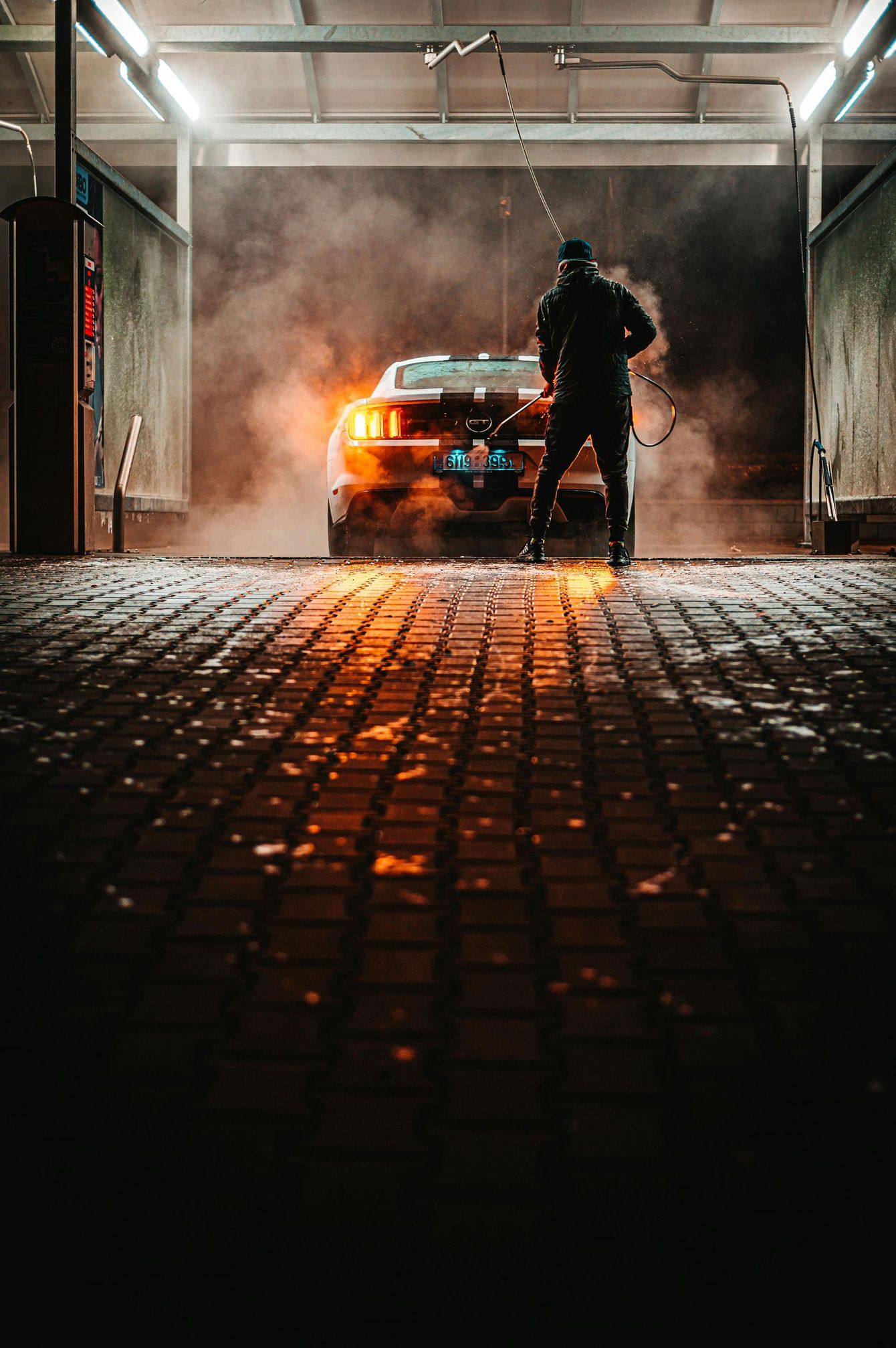 Person washing a car in a carwash, with steam and warm lighting reflecting on the cobblestone floor.