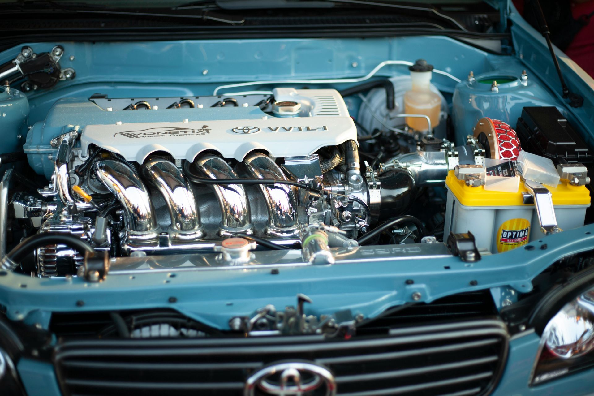 Blue car engine bay with polished components. Gold battery, silver pipes, and black grille visible.