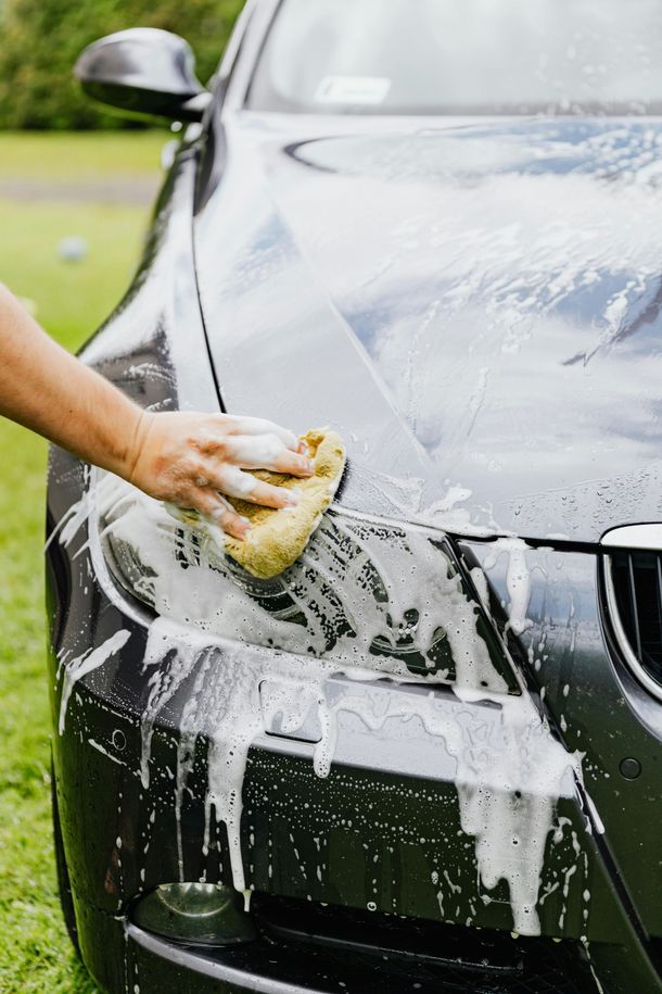 Person washing a dark-colored car with a soapy sponge outdoors on grass.