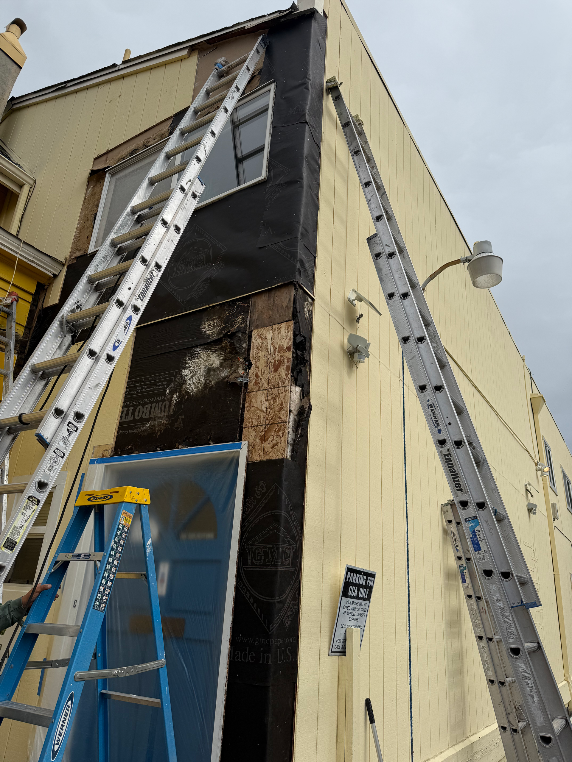Ladders on building exterior during construction; dark moisture barrier, light siding, blue door.