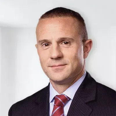 Man in a suit and tie, smiling, against a blurred white background.
