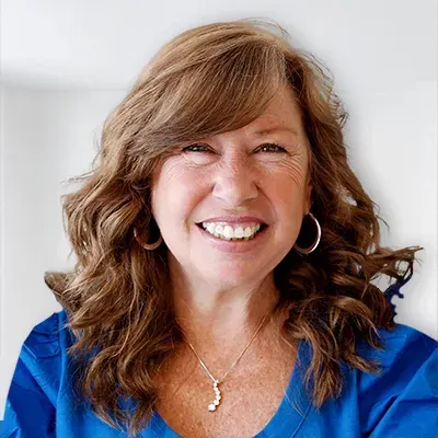 Woman with wavy auburn hair smiles, wearing a blue top, hoop earrings, and a pendant necklace.