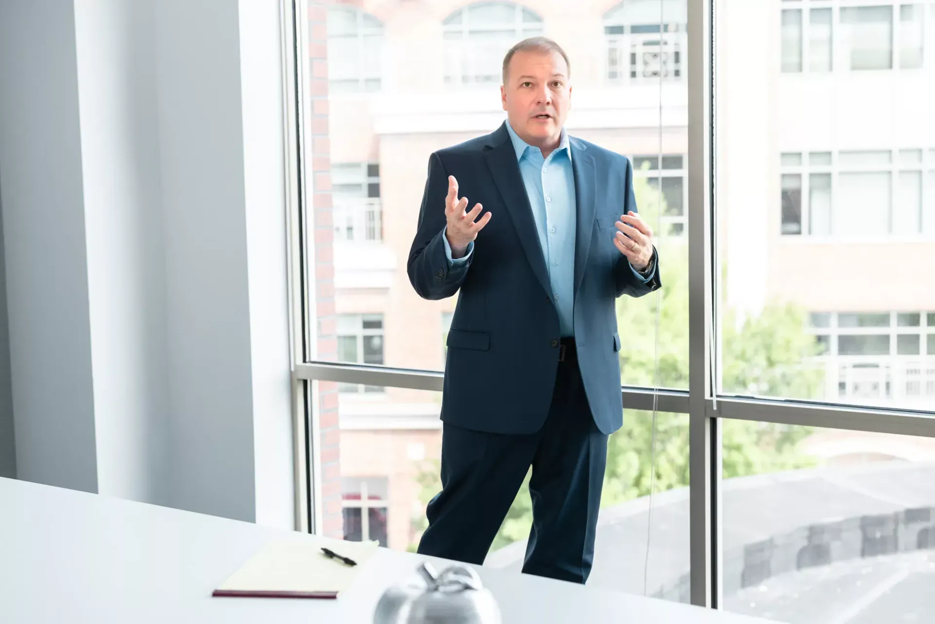 Man in blue suit gestures, standing near a window in a modern office, possibly giving a presentation.
