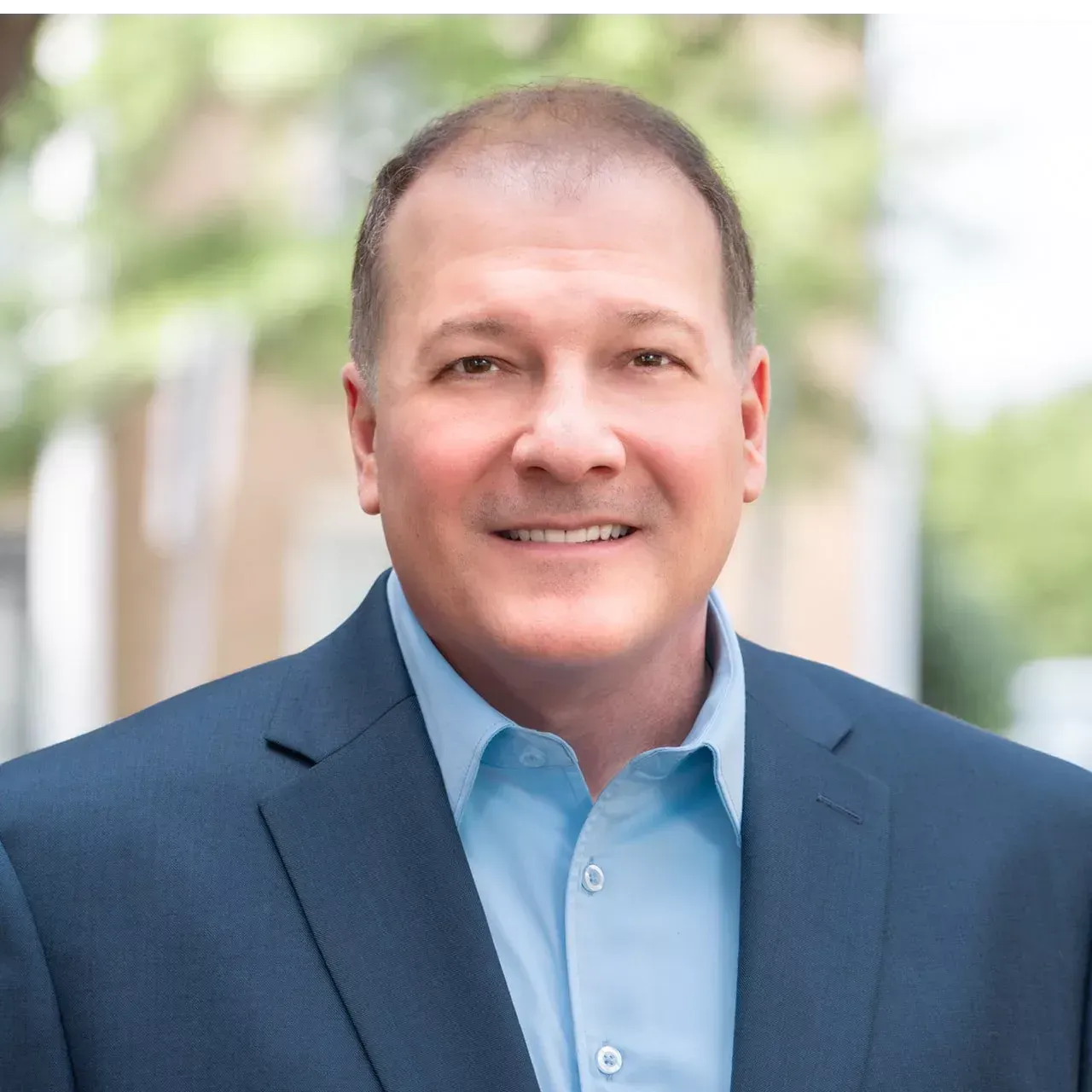 Man in blue suit and shirt smiles at the camera, blurred outdoor background.