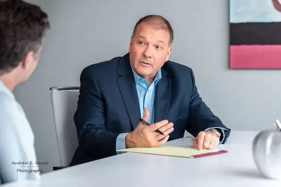 Man in suit gestures, speaking to another person at a table, likely in an office setting.