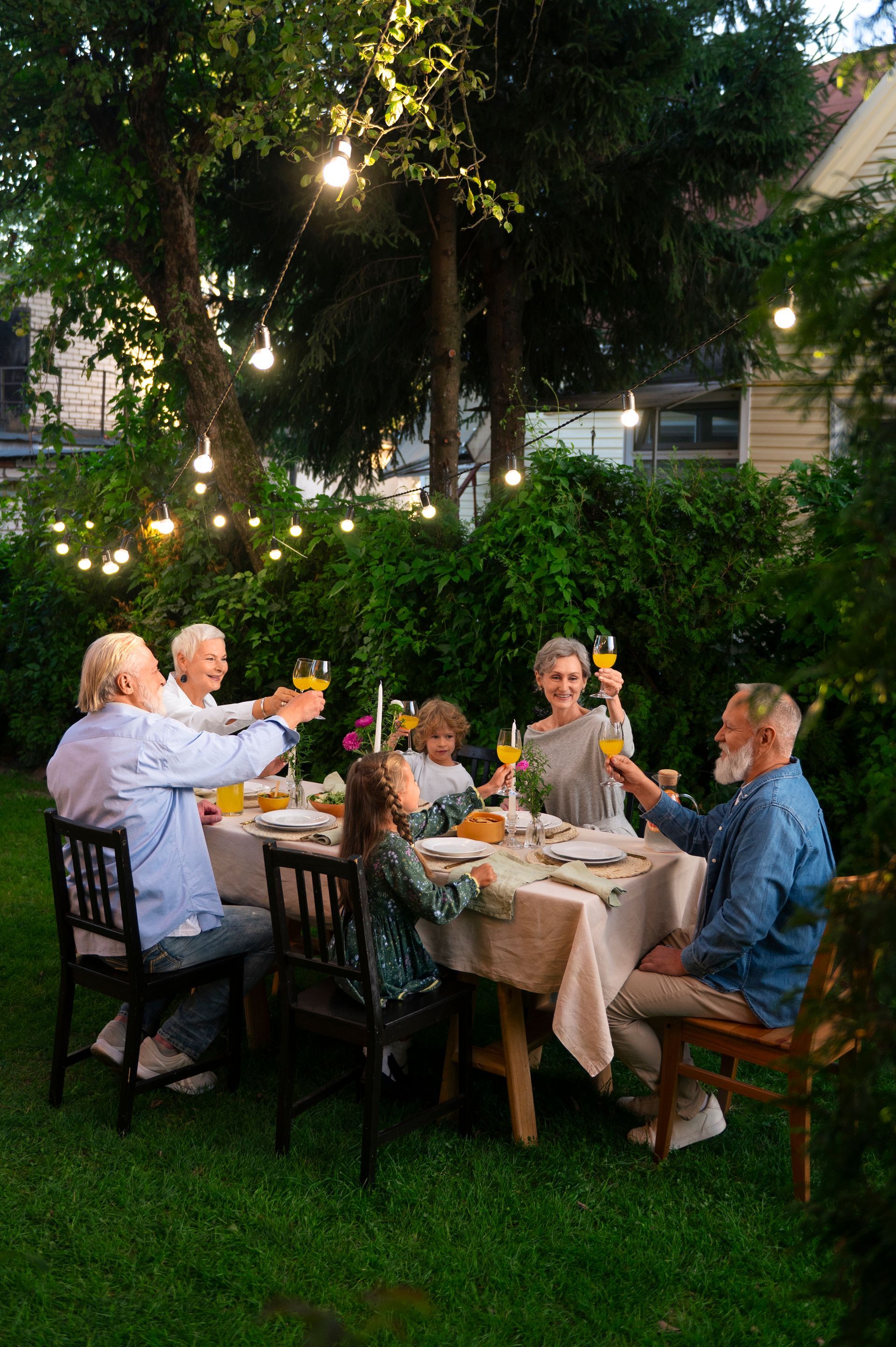 People toasting drinks around a table set for dinner outdoors, lit by string lights.