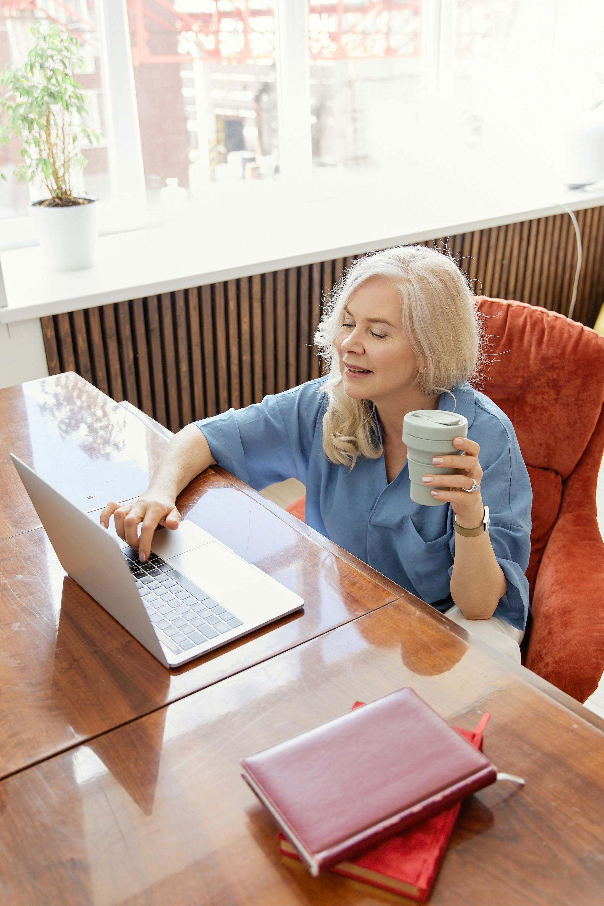 Woman sits at a table, using a laptop, holding a mug. Books on the table. Sunlight streams in.