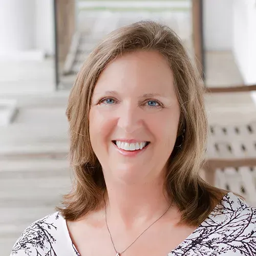 Woman with brown hair smiles, wearing a patterned top, set against a blurred background of stairs.