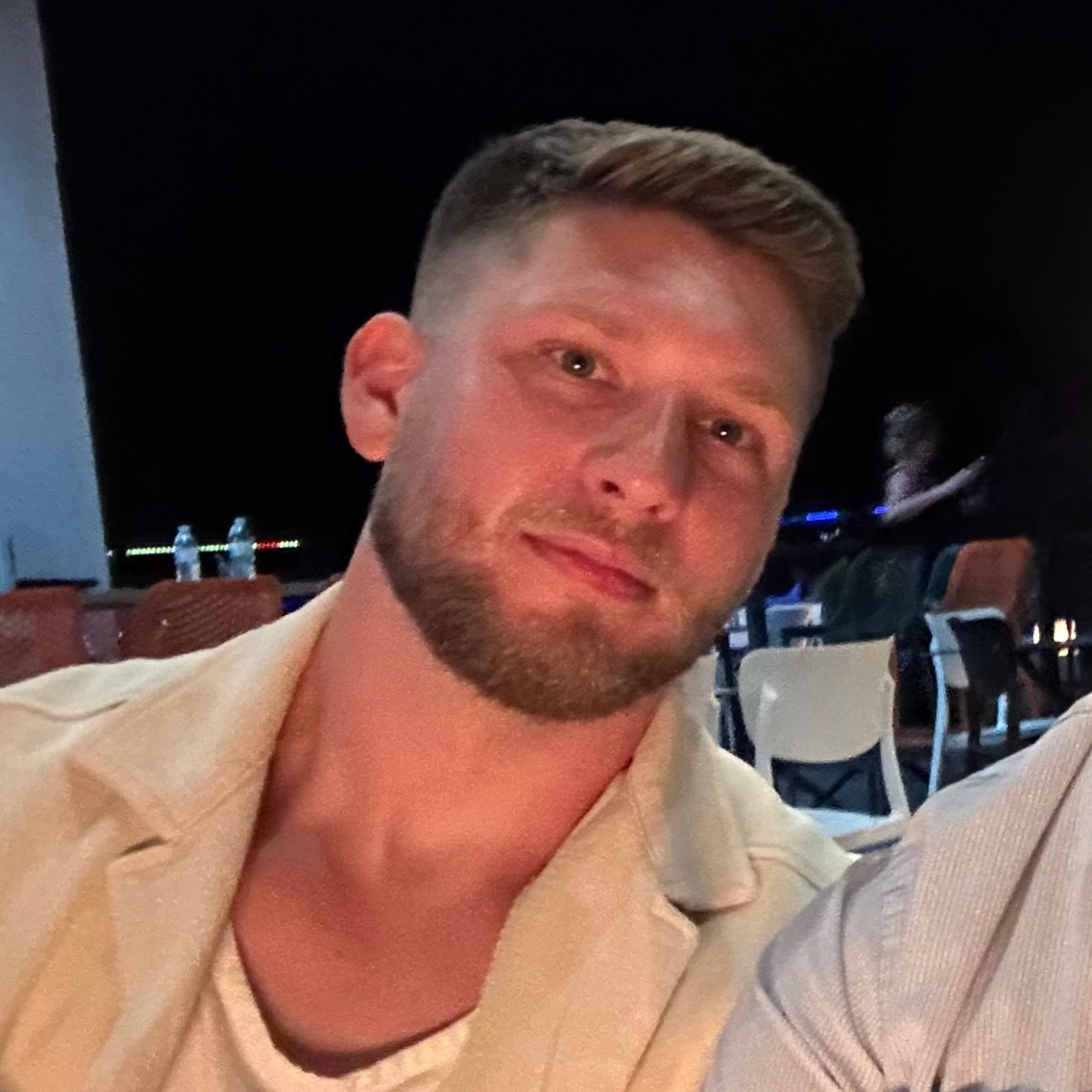 A man with a beard is wearing a white shirt and a tan jacket.