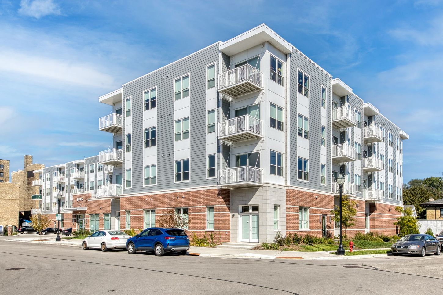 Modern apartment building with red brick base and gray siding. Cars parked on street.