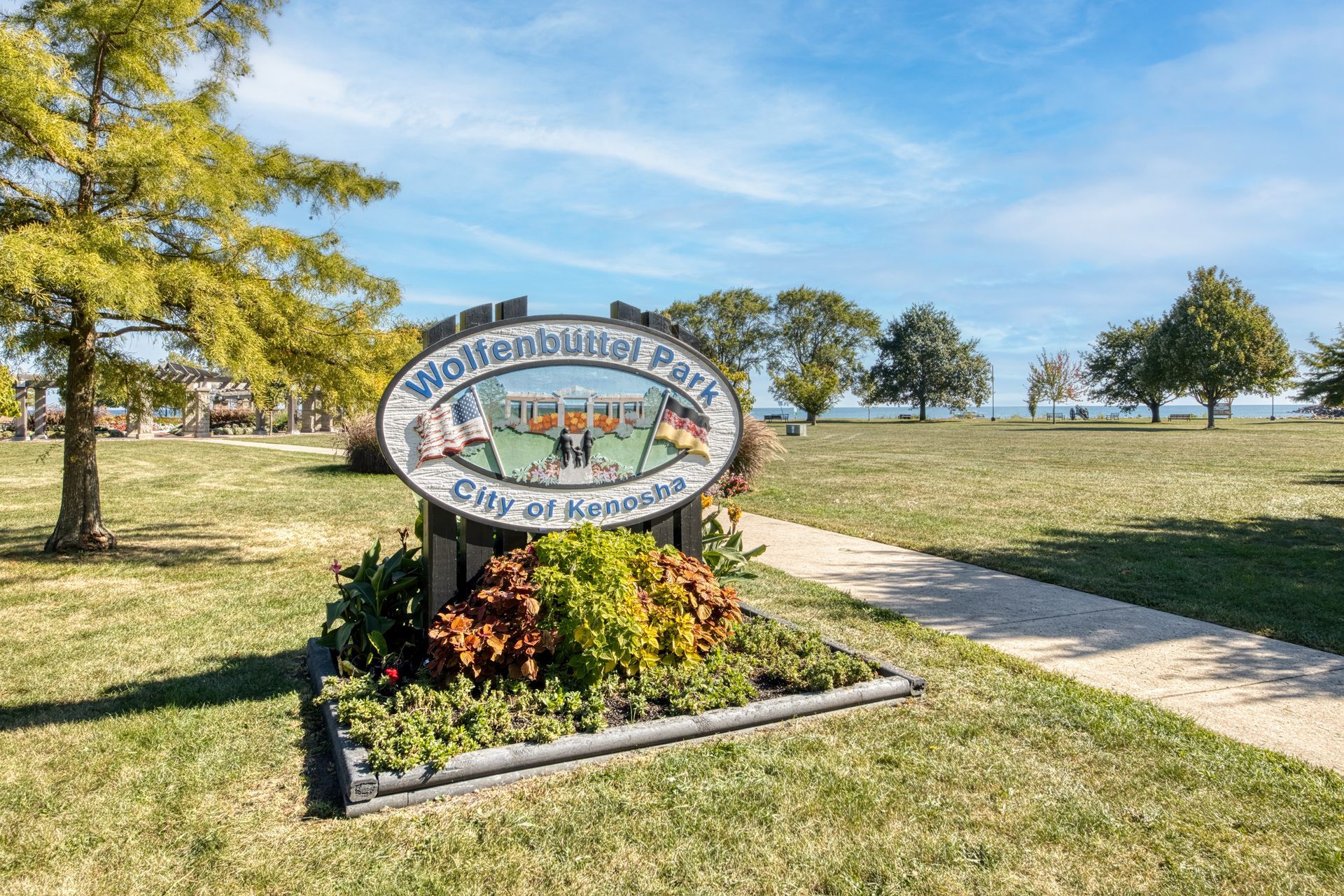 Sign for Wolfenbuttel Park, City of Kenosha, in a grassy park, with trees and a lake visible.