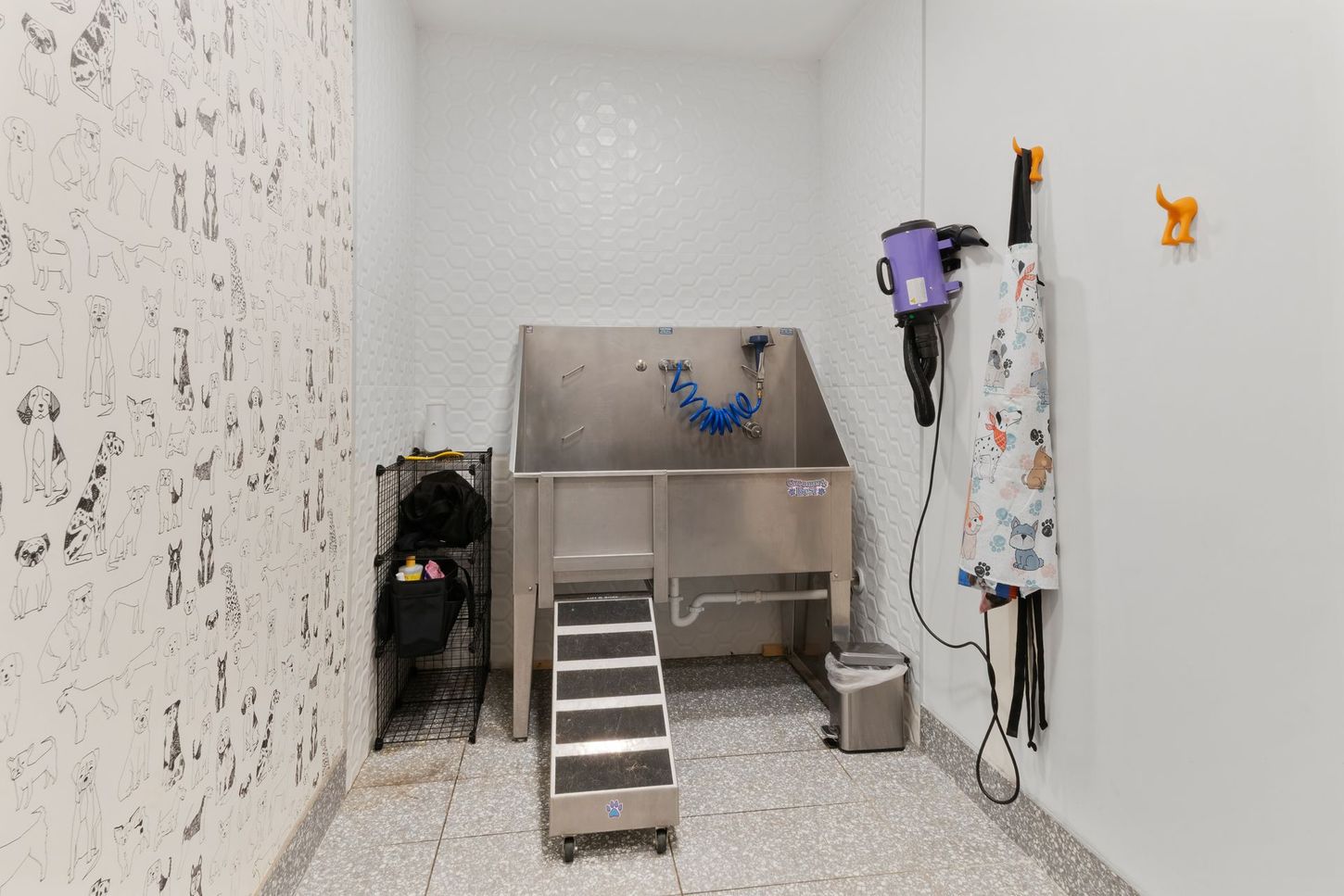 Dog grooming station with stainless steel tub, ramp, and dryer in a white-tiled room.