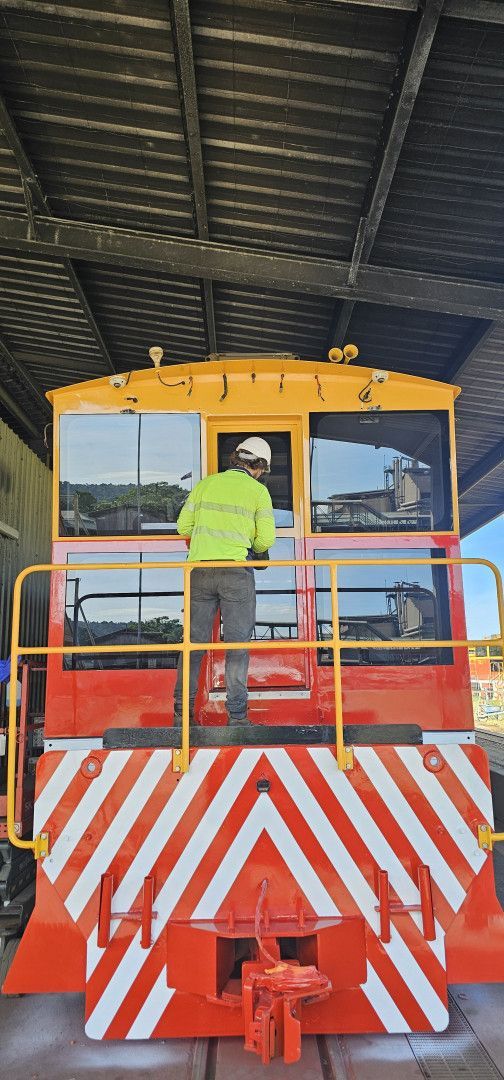Yellow locomotive front with red-and-white striped bumper under a covered shed, a worker standing on the platform