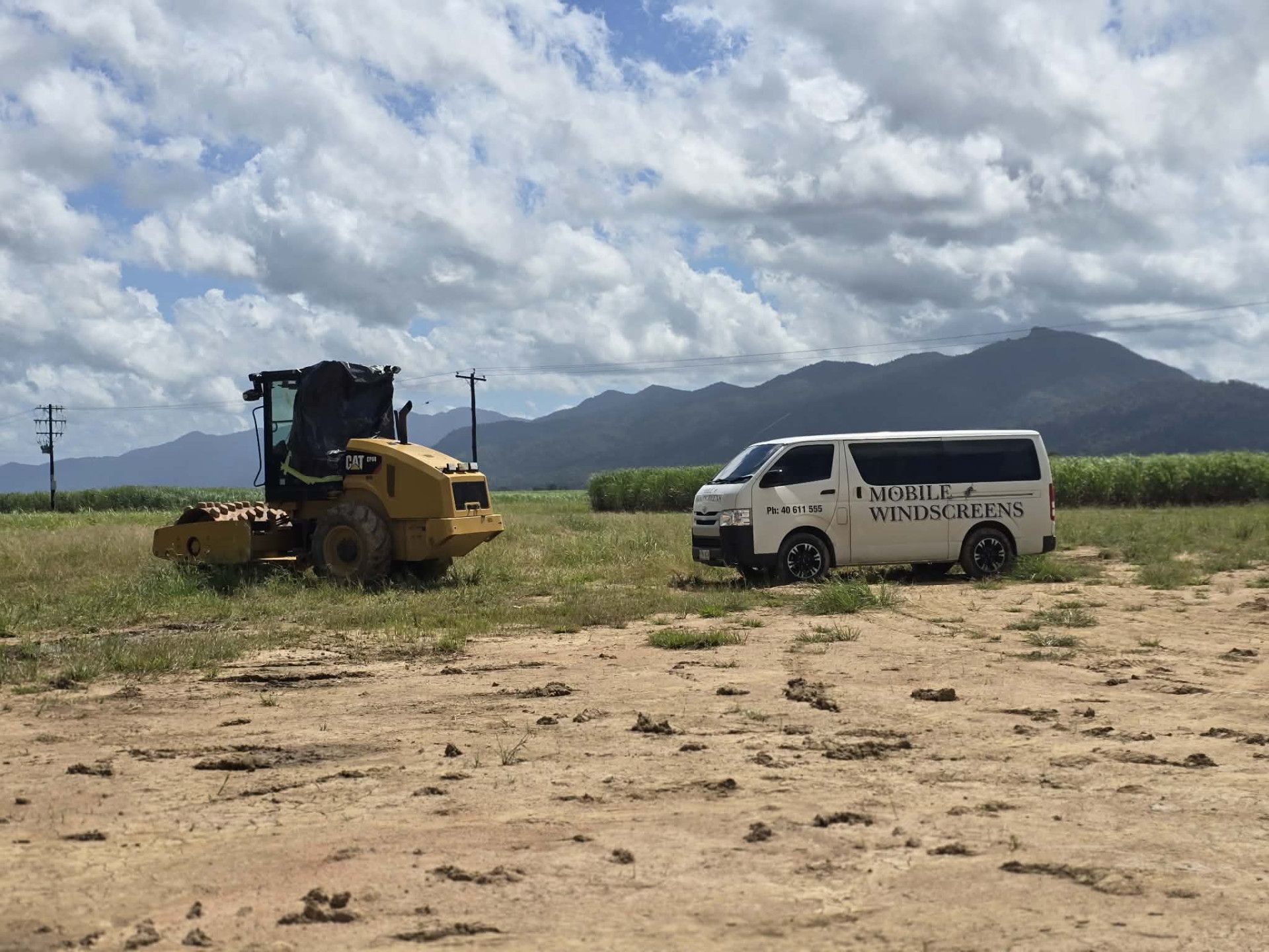 Yellow construction machine and white service van in a field with mountains and cloudy sky.