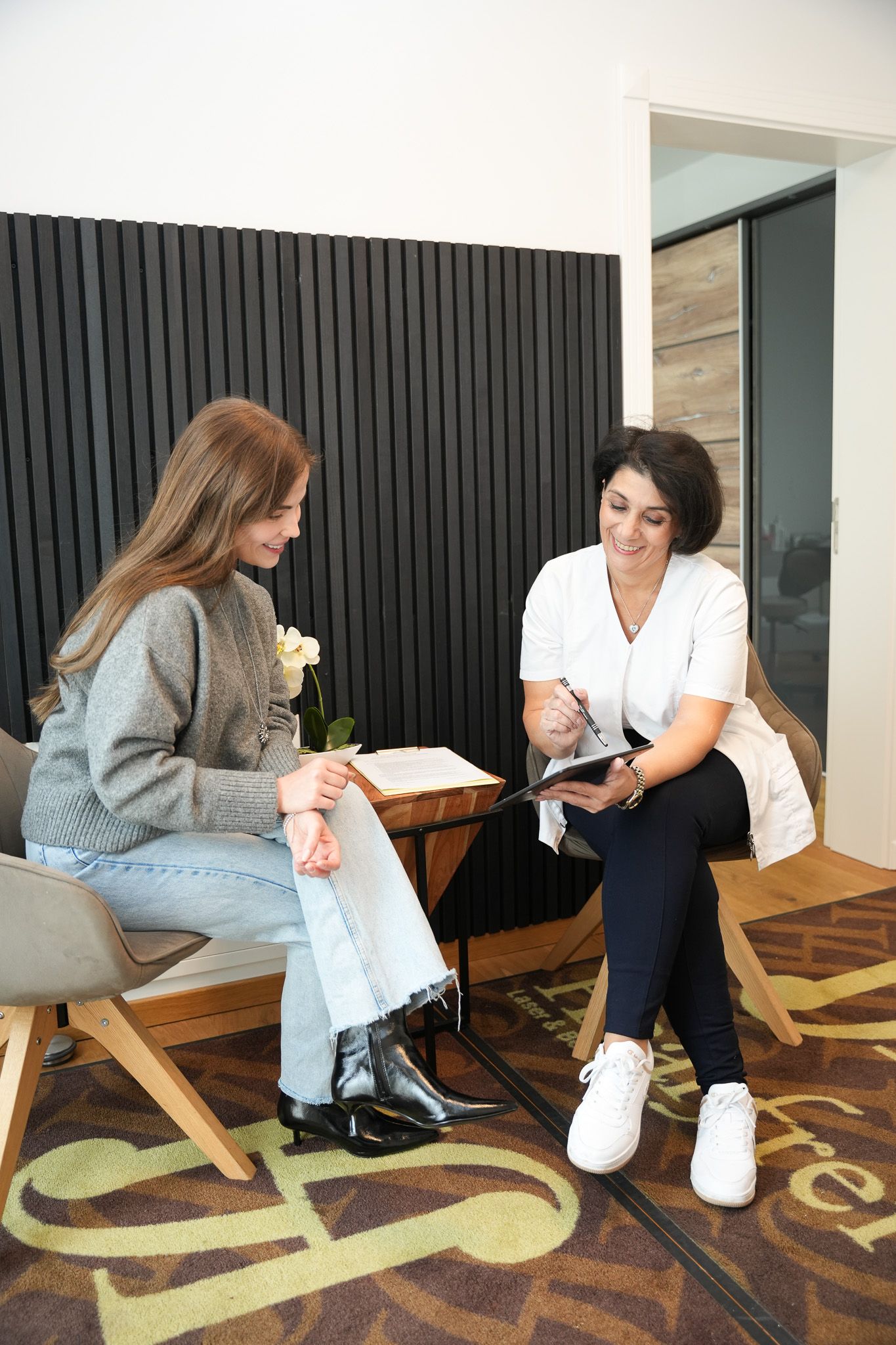 Eine Frau berät sich mit einer Fachkraft in einem modernen Büro. Beide sitzen, die Fachkraft hält ein Klemmbrett.