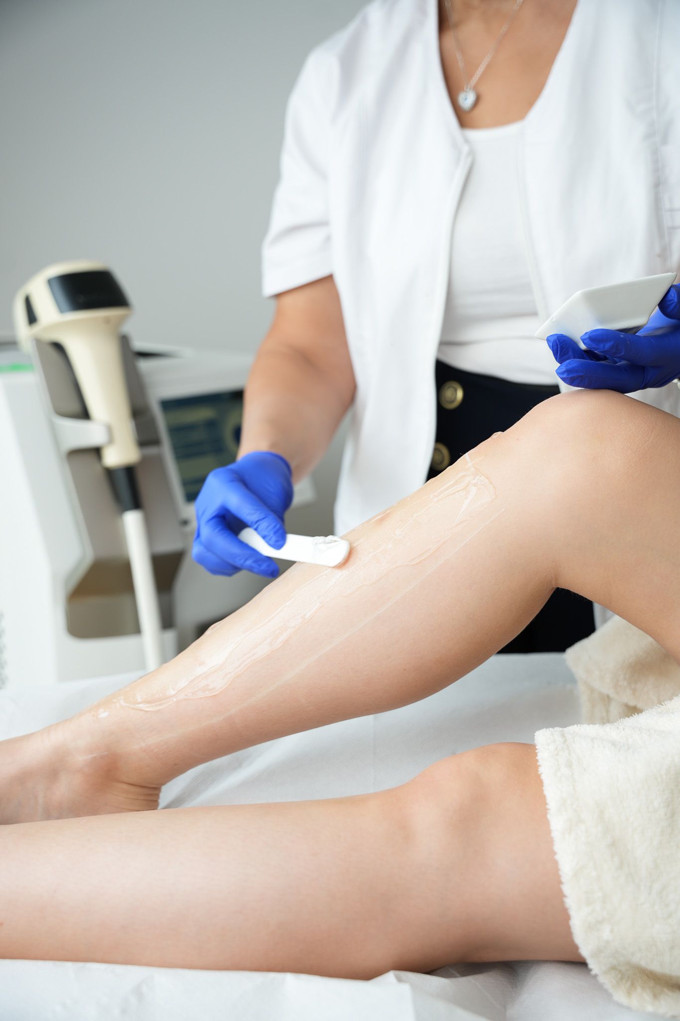Eine Person trägt blaues Gel auf ein Bein auf; im Hintergrund ist ein Laser-Haarentfernungsgerät zu sehen.