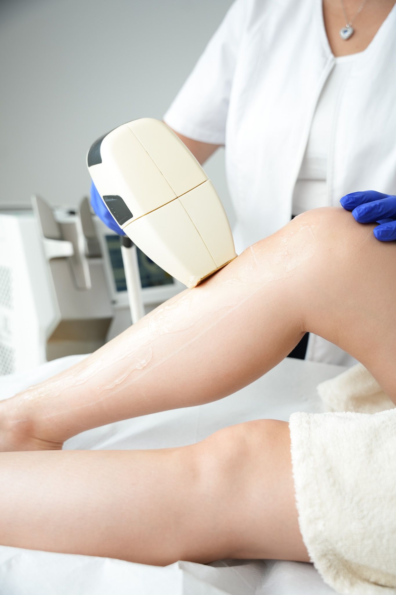 Eine Person mit blauen Handschuhen benutzt in einer klinischen Umgebung ein Laser-Haarentfernungsgerät an einem Bein.