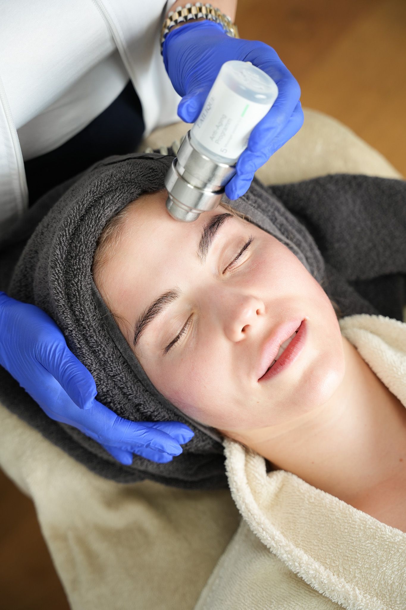 Frau erhält Gesichtsbehandlung; Therapeutin mit blauen Handschuhen hält ein Gerät an ihre Stirn.