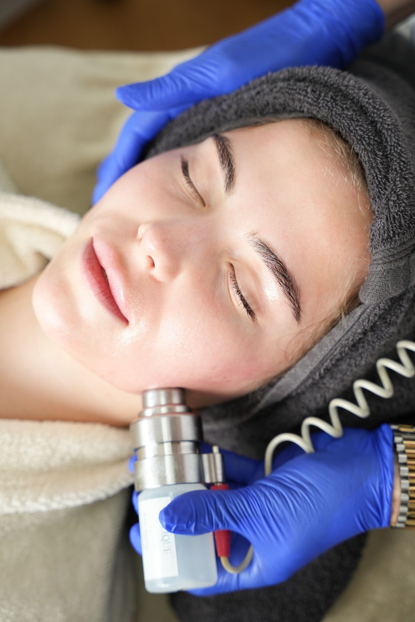 Frau bei Gesichtsbehandlung, Hände in blauen Handschuhen halten ein Gerät an ihren Kiefer, Augen geschlossen.