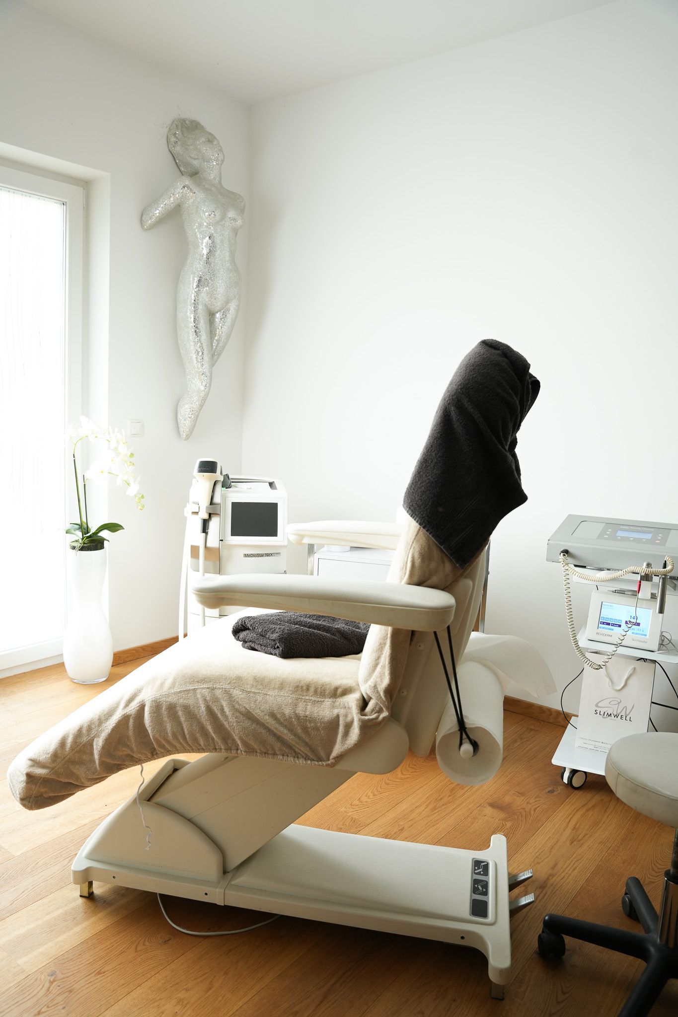 Ein mit Handtüchern bedeckter Behandlungsstuhl in einer Arztpraxis. Im Hintergrund medizinische Geräte und eine Skulptur.