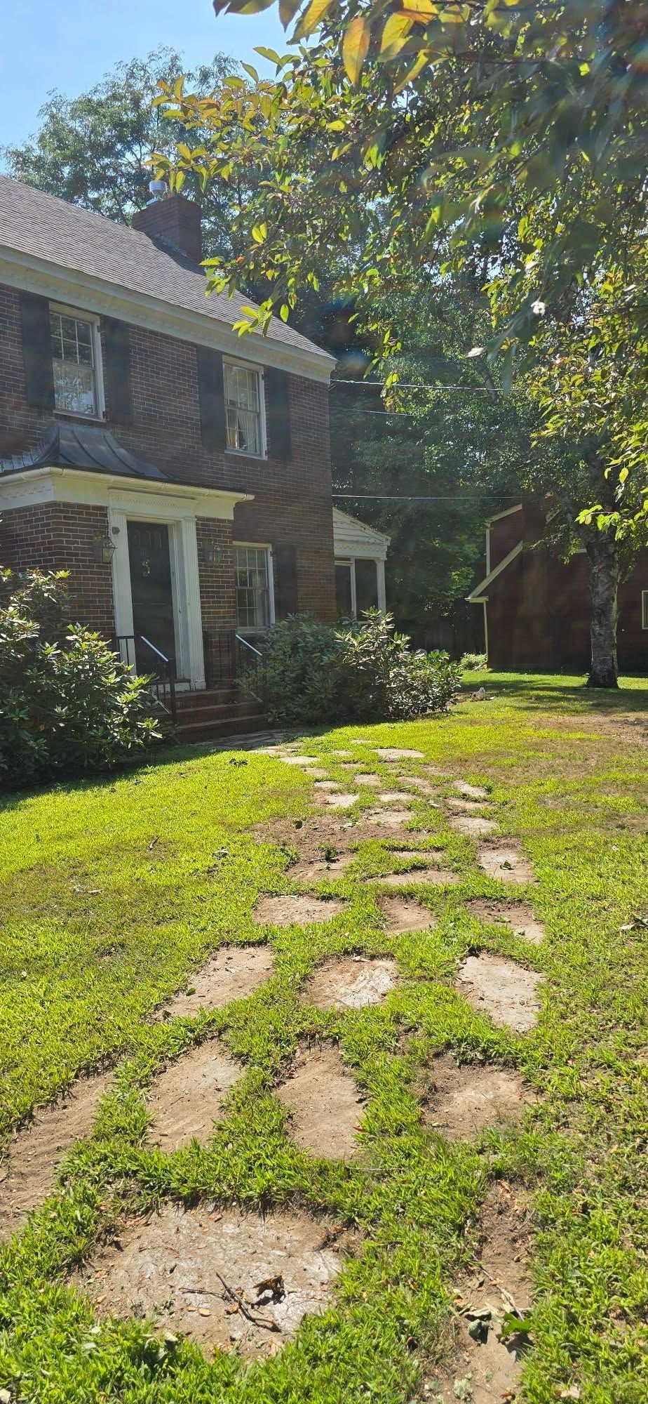 Stone pathway leads to a brick house with white porch and shutters, surrounded by green grass and trees.