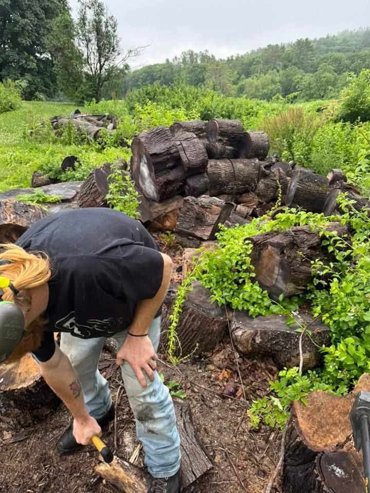 A person wearing safety glasses uses a hand tool to work on logs outside; green foliage surrounds them and stacked logs.
