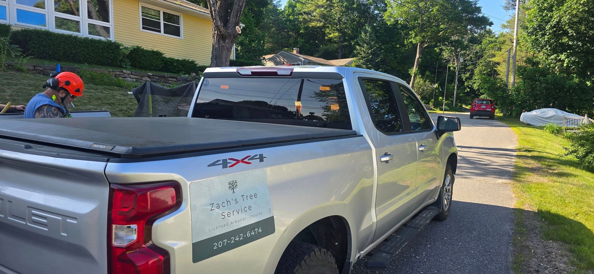 Silver pickup truck parked on gravel driveway, worker in background, yellow house.