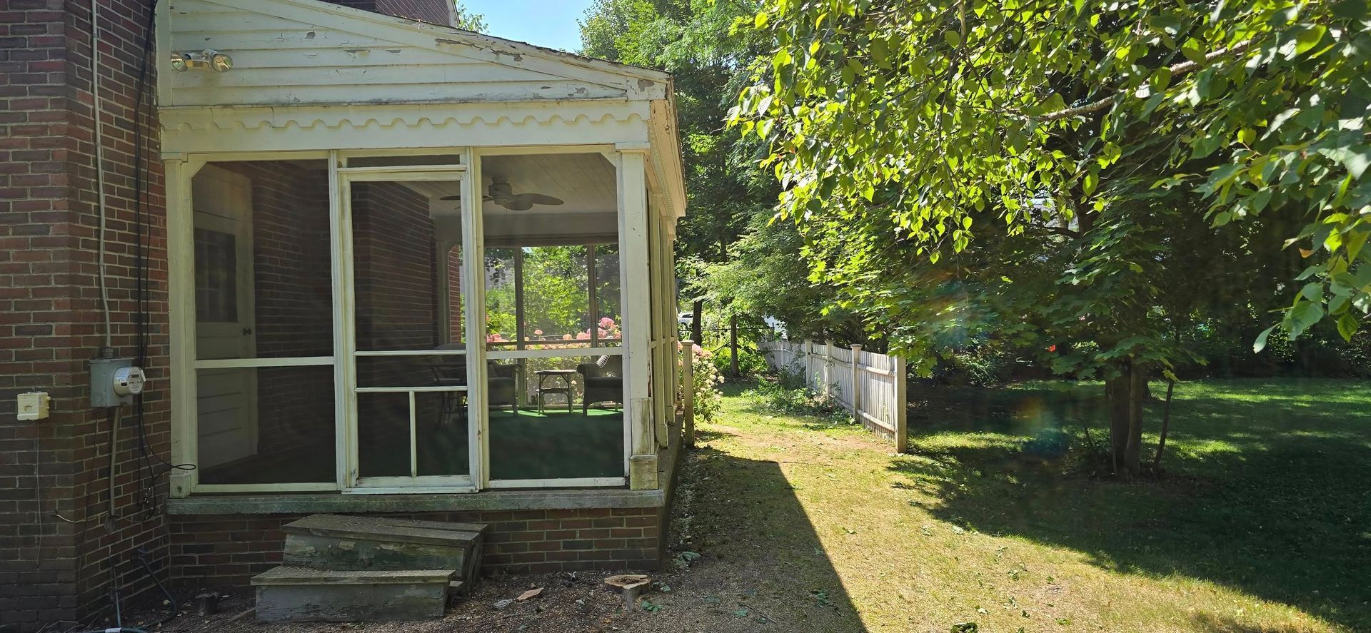 A screened porch on a brick building with a green lawn and a tree. A white fence is visible in the background.