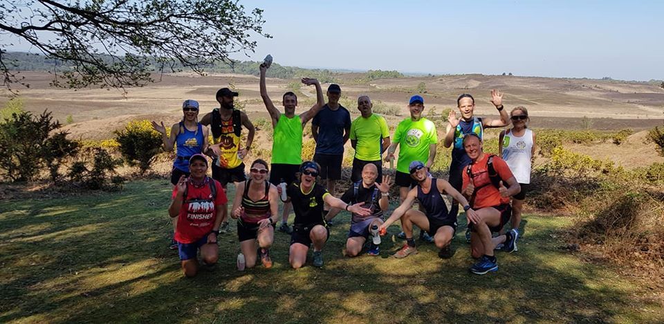 Group of runners posing on a grassy hilltop. They are waving at the camera, with a landscape view in the background. Sunny day.