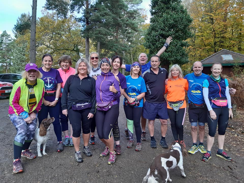 Group of runners posing outdoors with two dogs. Forest setting, some wearing running gear and colorful tops.