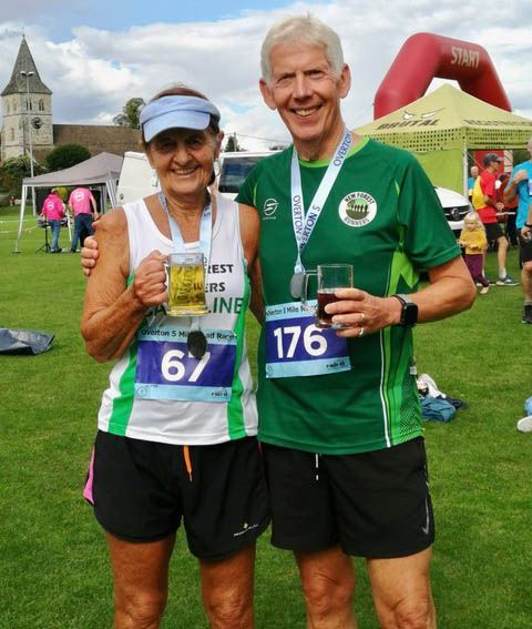 Two runners at an event, wearing race bibs and holding drinks, smiling. Outdoor setting with event tent.