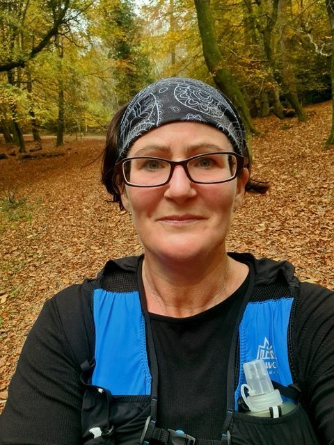 Woman in glasses and headband smiles in a wooded area, wearing a running vest.