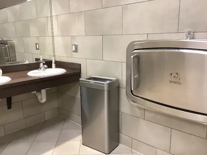 Public restroom with a changing station, sink, mirror, and a stainless steel trash can.