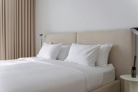 A modern bedroom with a bed, white linens, beige headboard, neutral curtains, and bedside table.