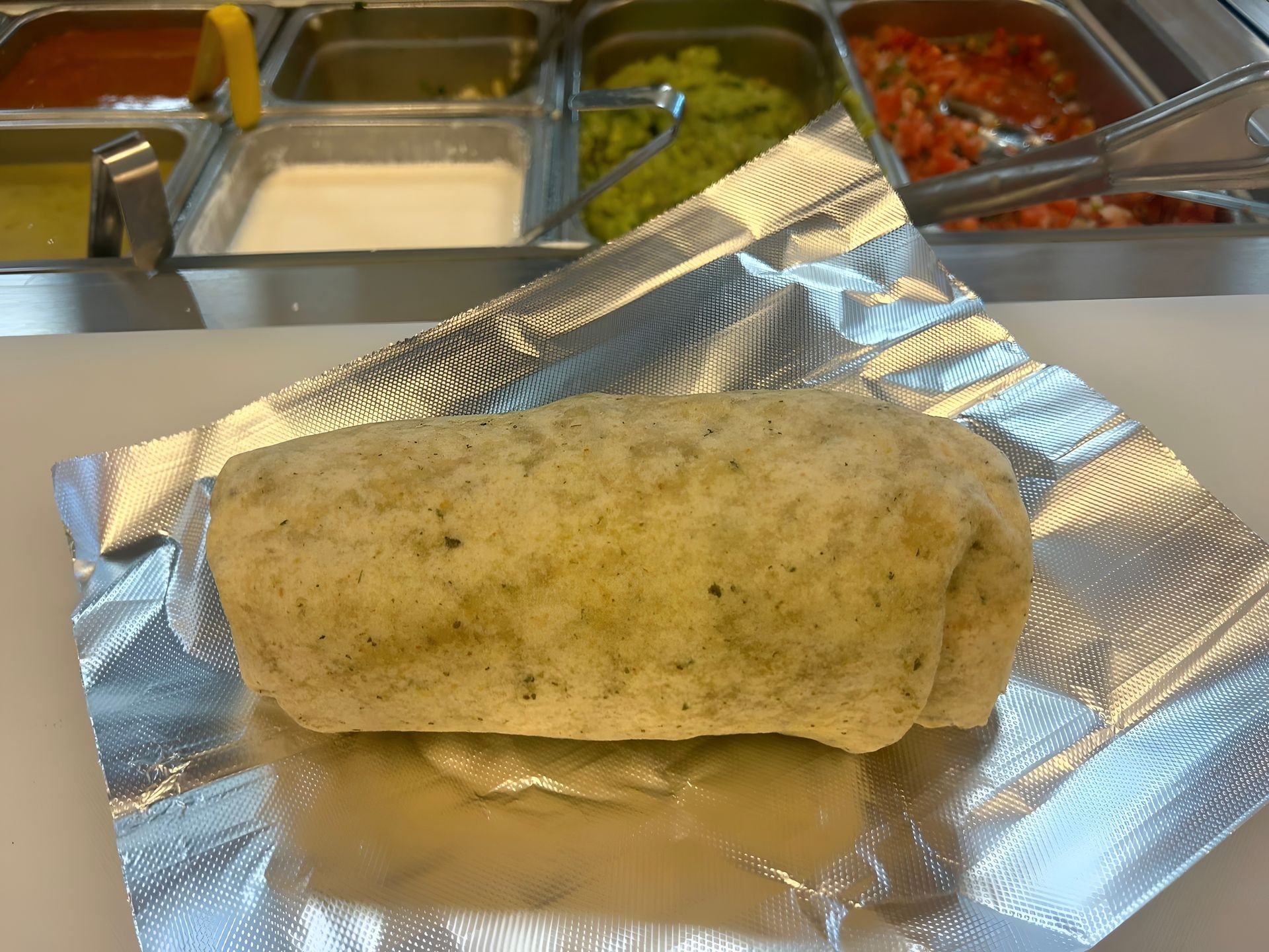 Burrito in foil wrapper; toppings in the background.