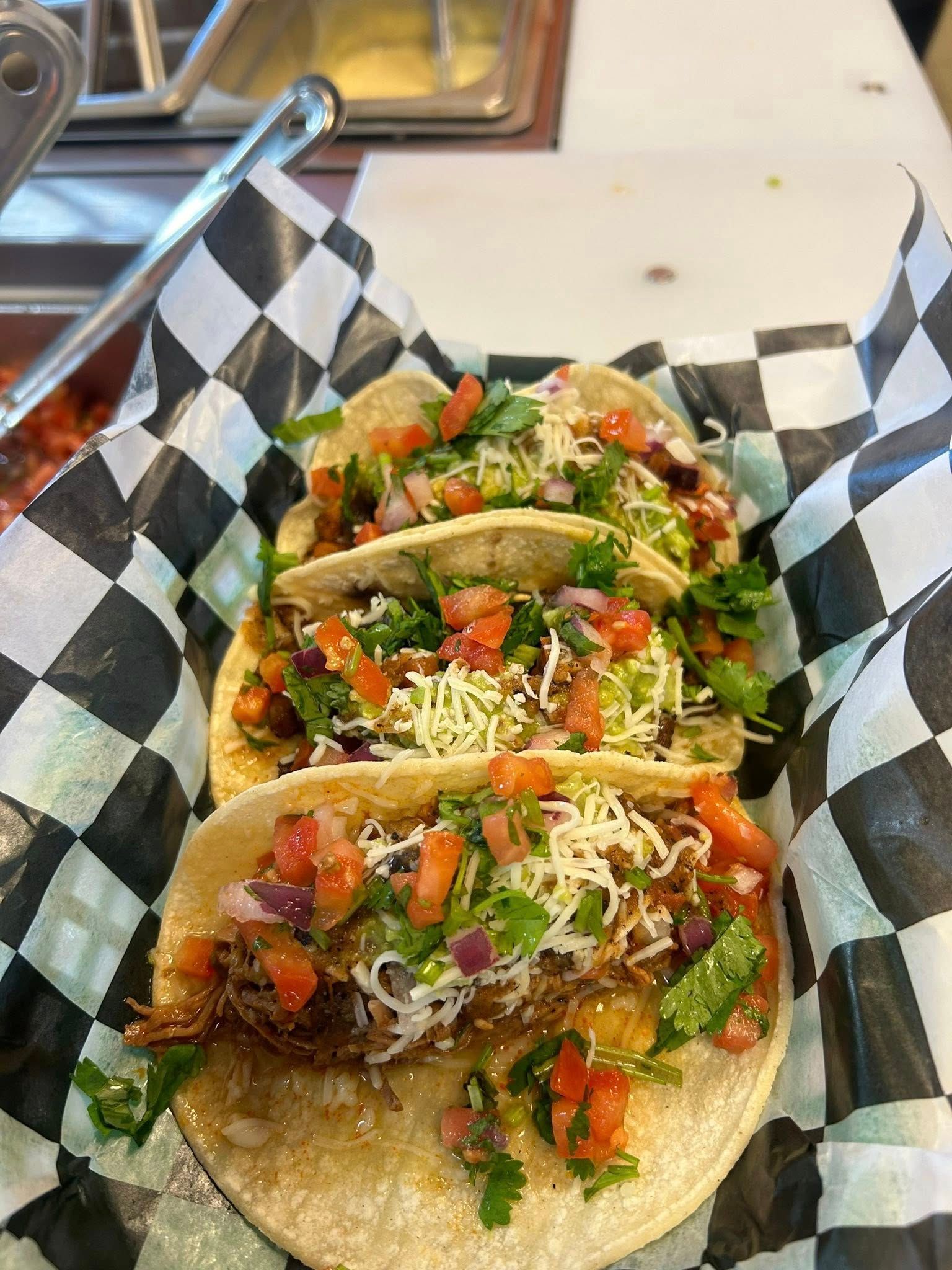 Three tacos in a checkered paper-lined basket, topped with pico de gallo, cilantro, and shredded cheese.