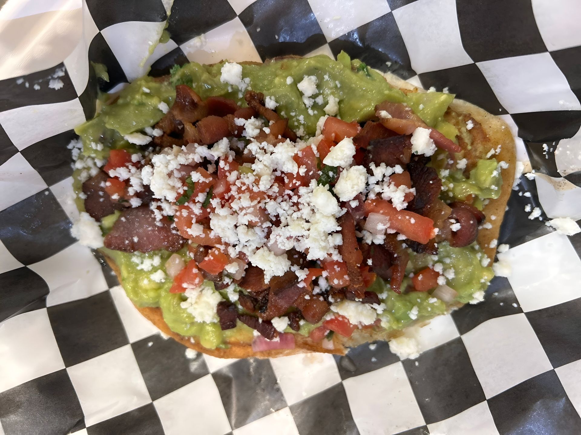 Tostada topped with guacamole, bacon, tomatoes, and crumbled cheese on checkered paper.