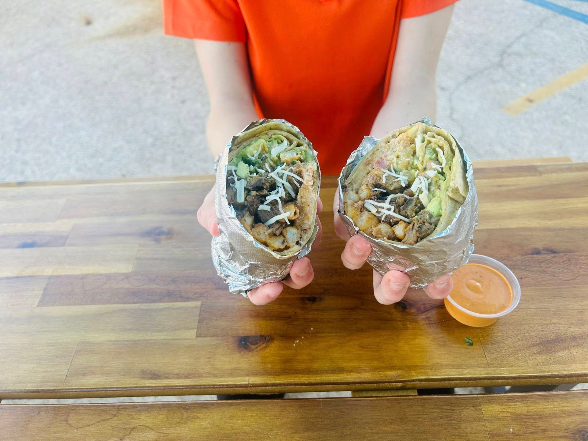 Person holding two halves of a burrito, wrapped in foil, over a wooden table, next to a small cup of orange sauce.