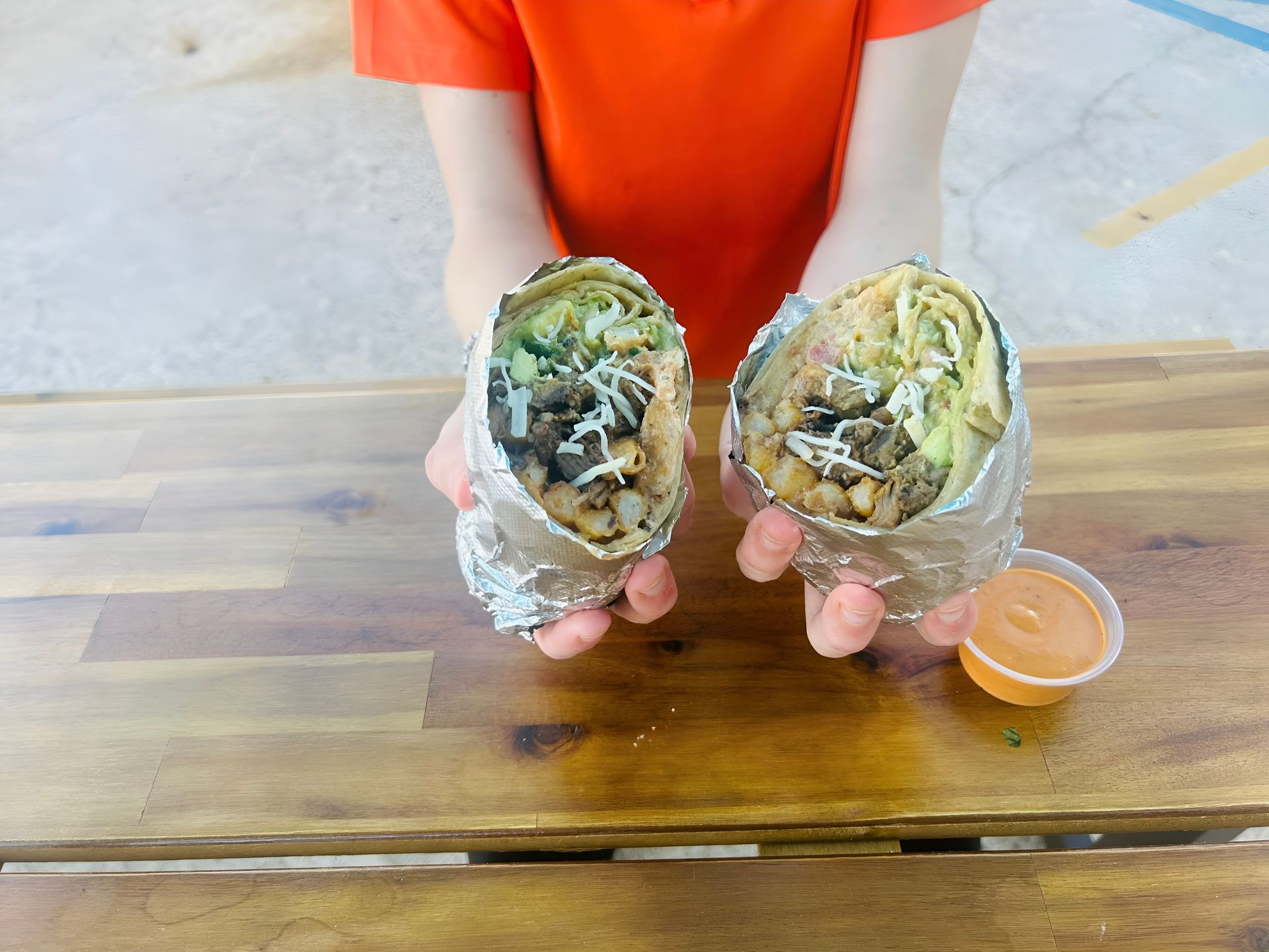 Person holding two halves of a burrito wrapped in foil, on a wooden table, with dipping sauce.