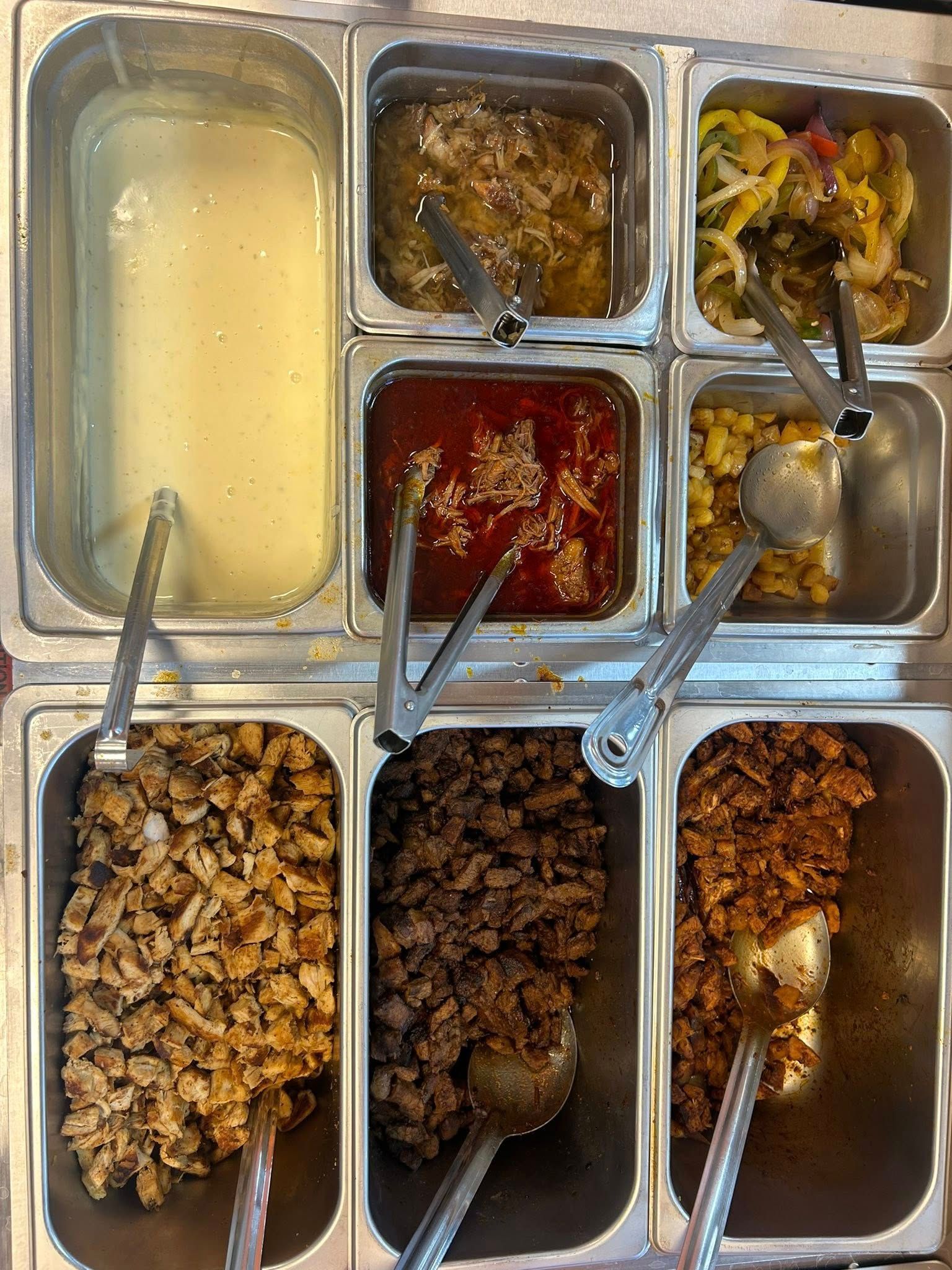 Food bar with various toppings and fillings in metal containers, with serving utensils.