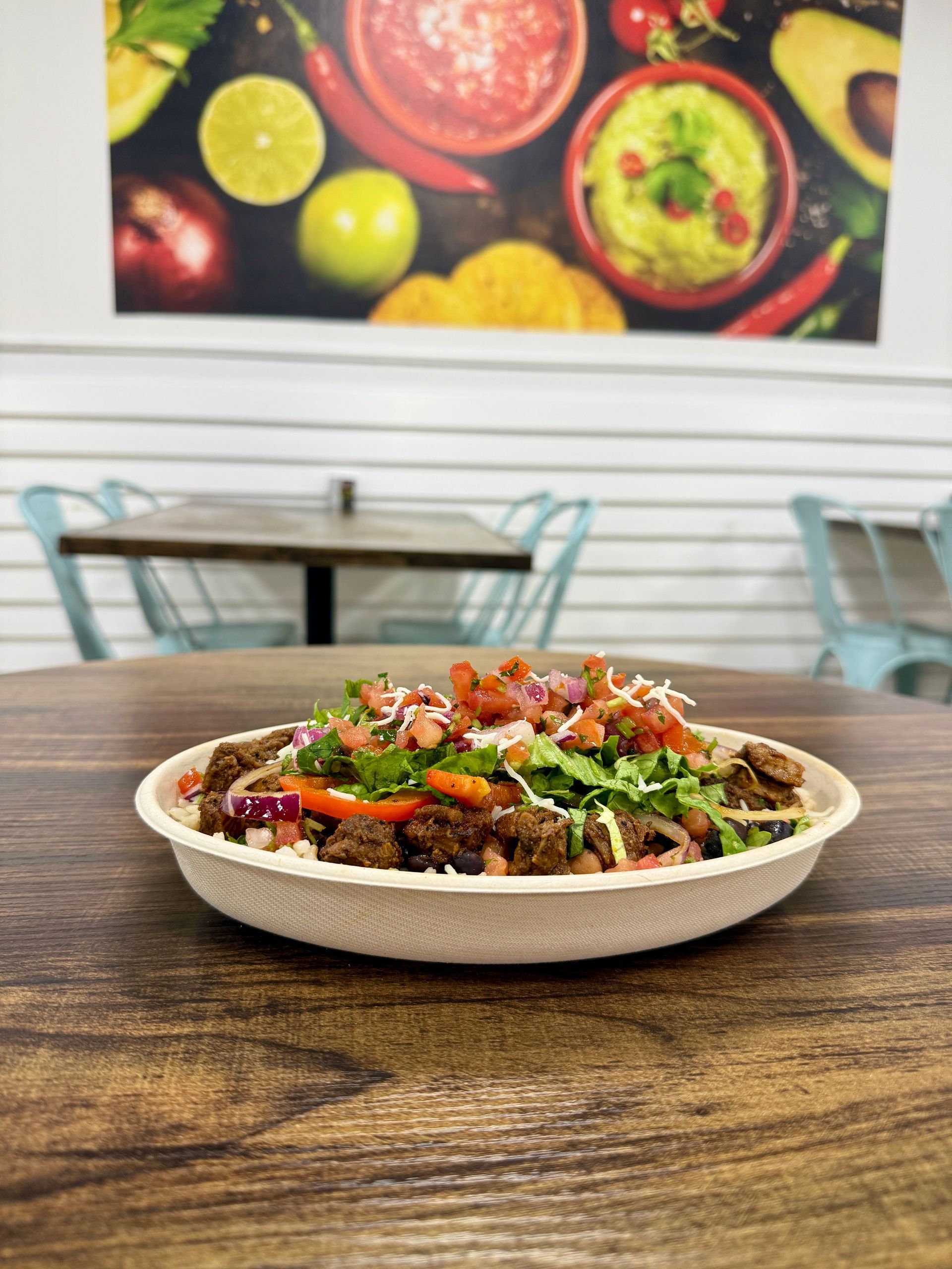 Close-up of a bowl filled with food on a wooden table. The bowl has meat, vegetables, and toppings. Background has a colorful food image.