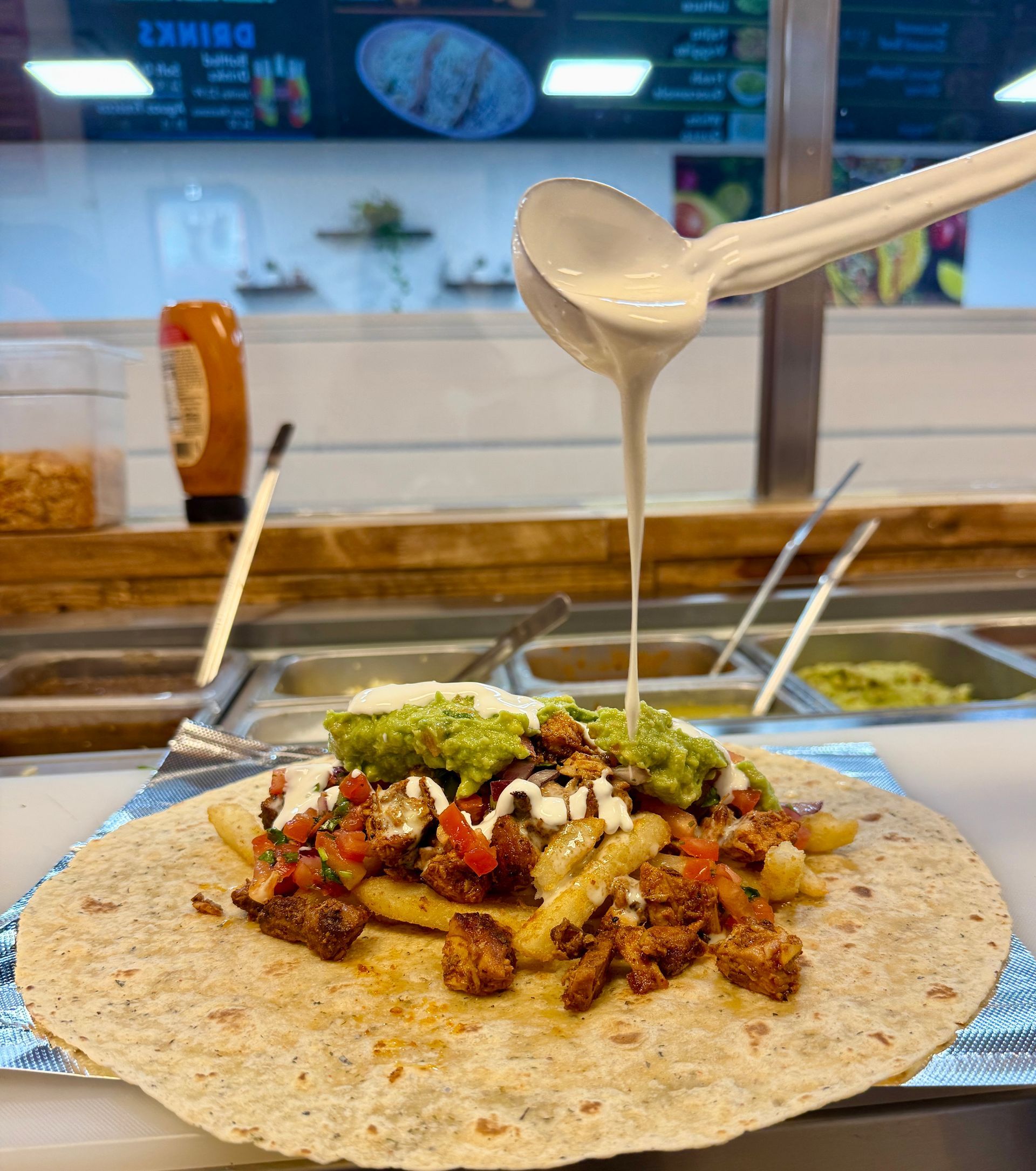 Burrito being drizzled with white sauce in a restaurant. It has fillings like meat, guacamole, and fries.