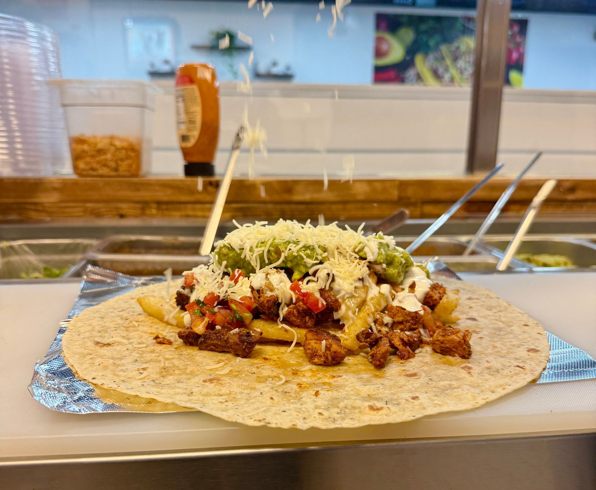 Burrito being assembled: tortilla base with meat, fries, cheese, and guacamole, with cheese being sprinkled on top.