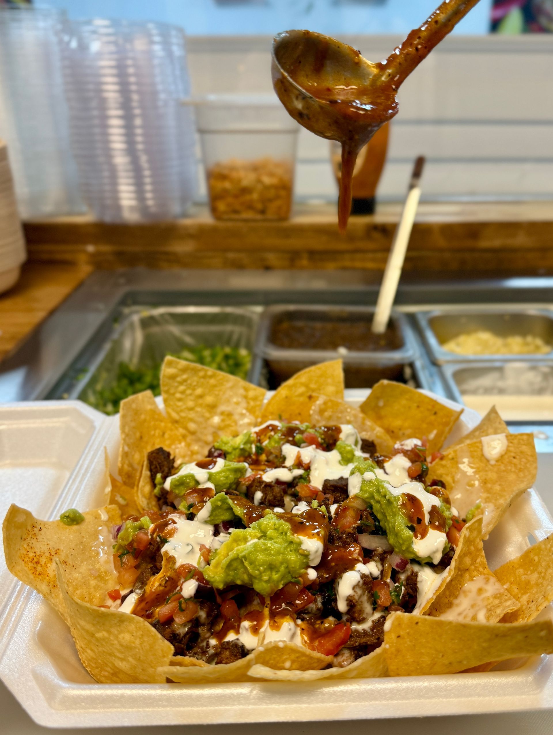 Nachos in a white container, topped with guacamole, sour cream, and salsa. Someone pours salsa from a ladle.